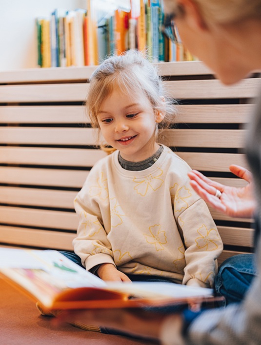 Small girl smiling at a book with a woman gesturing in the foreground